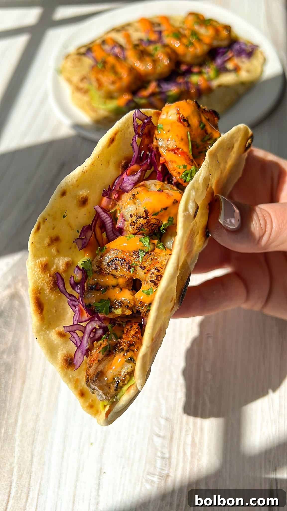 Quick and Delicious Shrimp Tacos 2 Close-up of a hand holding a taco with grilled shrimp, purple cabbage, and herbs in a soft tortilla. A similar loaded taco is blurred in the background on a white plate. Sunlight casts shadows on the wooden surface.