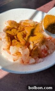 A white bowl filled with raw shrimp is being sprinkled with a generous amount of brown seasoning from a spoon. The shrimp are pale pink and plump, and the seasoning adds a textured, spicy-looking layer on top.