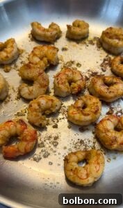 Shrimp seasoned with spices are cooking in a stainless steel pan. Some have a golden-brown sear, indicating they are nearly cooked. The shrimp are arranged randomly, with visible seasoning flakes scattered throughout the pan.