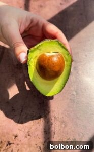 A hand holding a halved avocado with a visible pit, resting on a textured brown surface. Sunlight casts shadows on the surface, highlighting the avocado's green flesh.