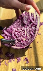 A hand holds half of a red cabbage on a wooden cutting board. A vegetable peeler is used to shave thin slices from the cabbage, revealing intricate patterns of purple and white layers.