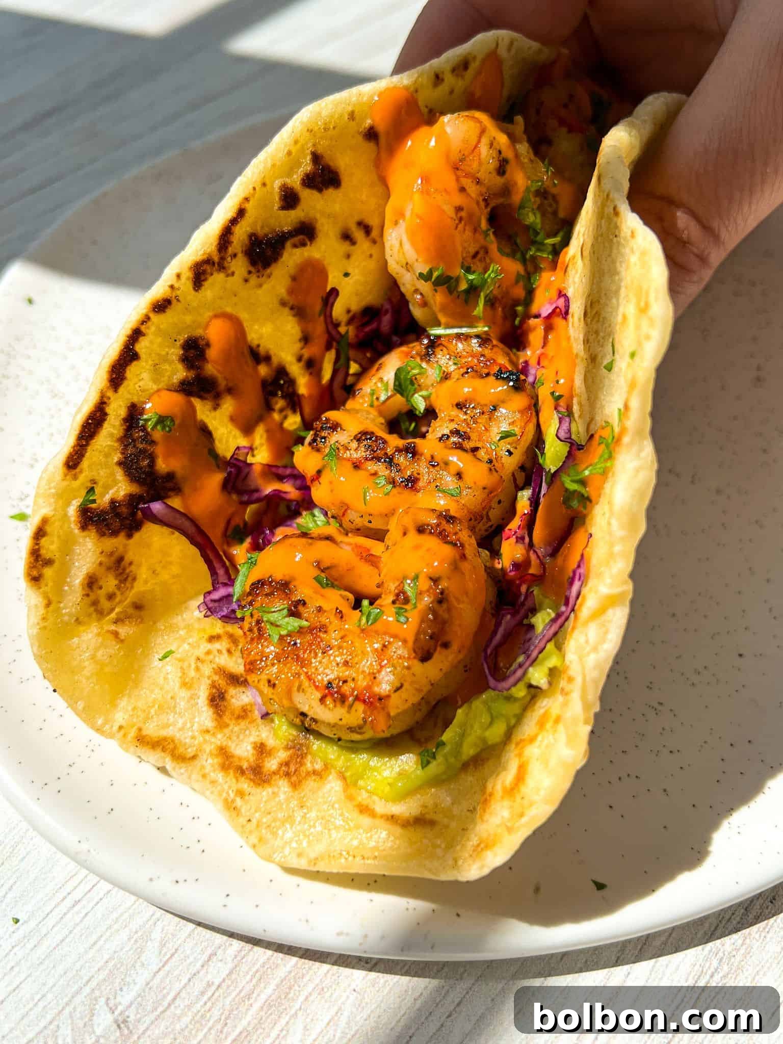 Quick and Delicious Shrimp Tacos 3 A hand holding a taco filled with grilled shrimp, shredded purple cabbage, and topped with an orange sauce, all wrapped in a lightly toasted tortilla, sitting on a white plate. Sunlight casts shadows on the surface.