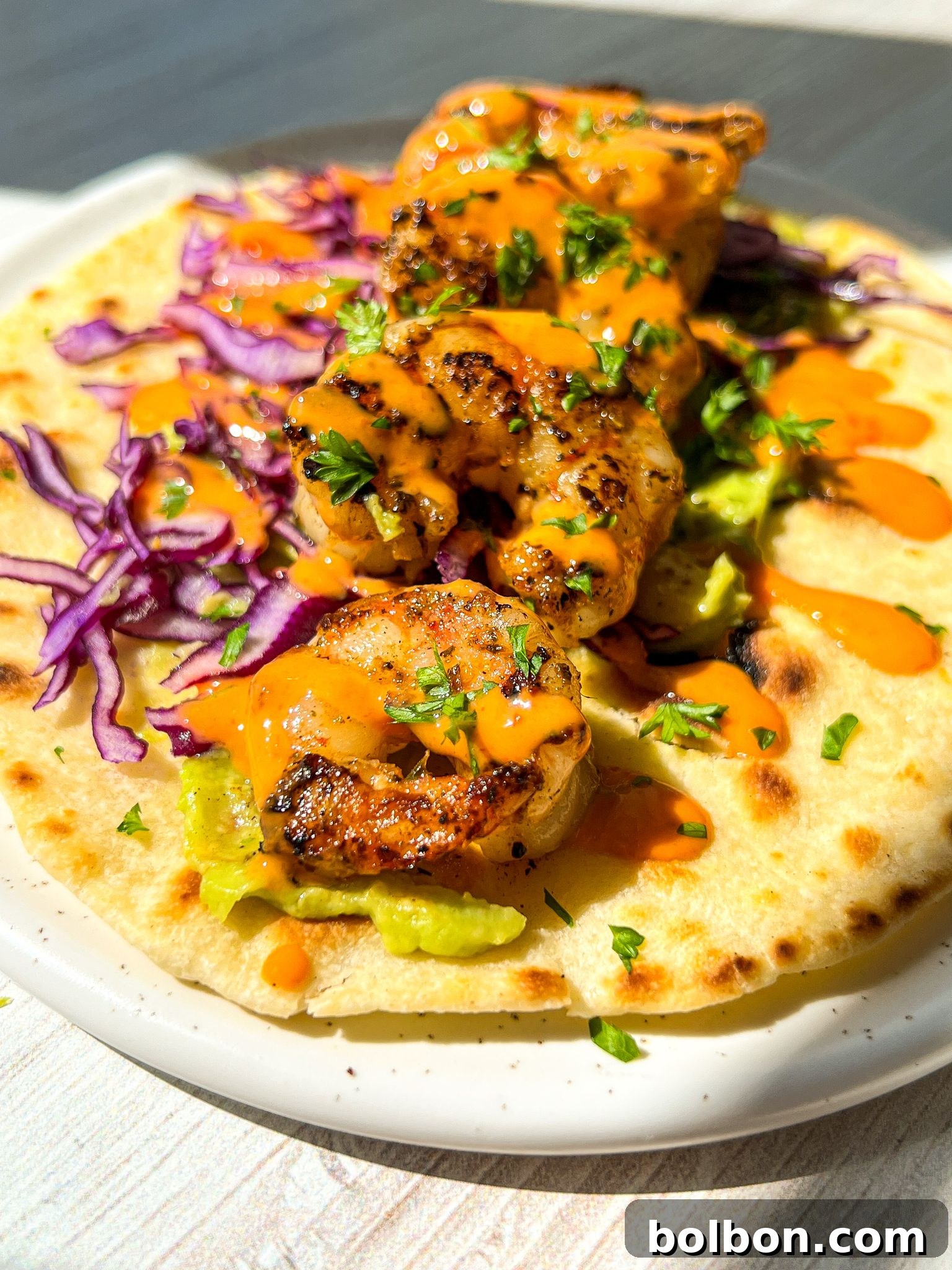 Quick and Delicious Shrimp Tacos 4 A grilled shrimp taco on a white plate, topped with vibrant red cabbage, avocado, and drizzled with orange sauce. The taco is garnished with chopped herbs, all set against a sunlit background.