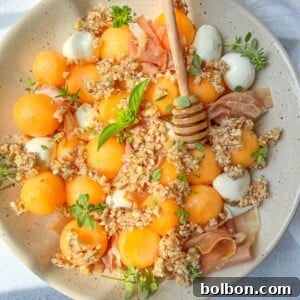 A speckled beige plate features prosciutto and melon balls, creamy cheese dollops, granola clusters, fresh herbs, and a honey dipper drizzled with honey for a savory-sweet delight.