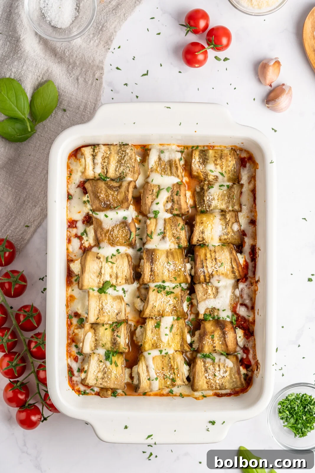 Savory Eggplant Spirals 2 A white rectangular baking dish filled with golden-brown eggplant rollatini, generously garnished with freshly chopped herbs. The setting includes vibrant cherry tomatoes, fragrant fresh basil leaves, aromatic garlic cloves, a small bowl of grated Parmesan cheese, a rustic wooden spoon, and a soft linen cloth, suggesting a rustic and fresh culinary experience.