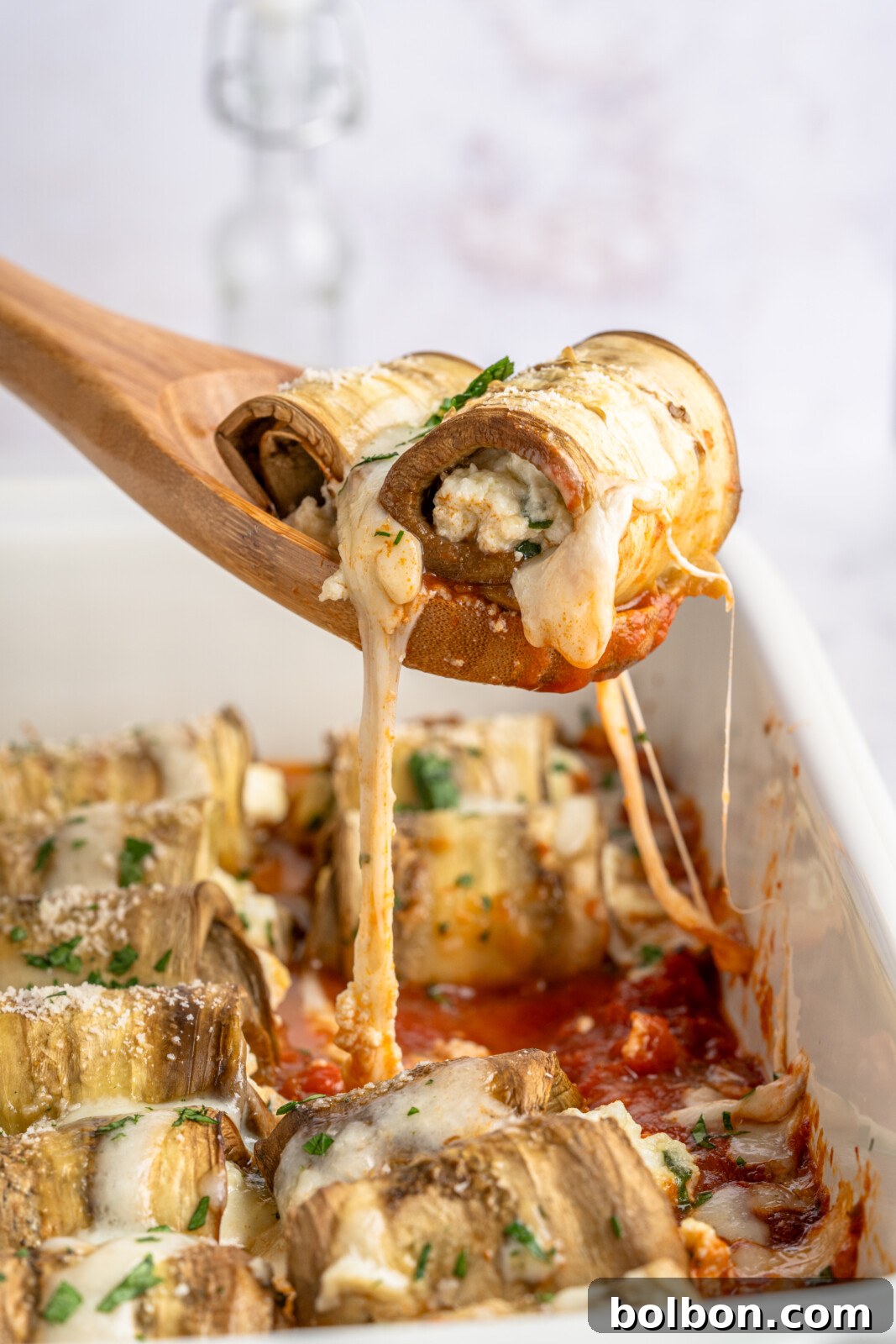 Savory Eggplant Spirals 3 A wooden spoon delicately lifts a serving of stuffed eggplant rollatini from a white baking dish. The rollatini glistens with melted cheese and is adorned with a sprinkle of chopped fresh herbs. The baking dish, still full, showcases more perfectly baked eggplant pieces nestled in a rich tomato sauce, all topped with a luscious layer of melted cheese, ready to be enjoyed.