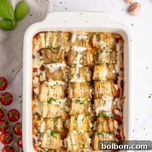Baked Eggplant Rolls (rollatini) in a white baking dish, topped with melted cheese and fresh herbs. The dish is surrounded by fresh cherry tomatoes, basil leaves, garlic cloves, a small pile of salt, and a gray napkin on a beautiful marble surface, emphasizing freshness and Italian tradition.