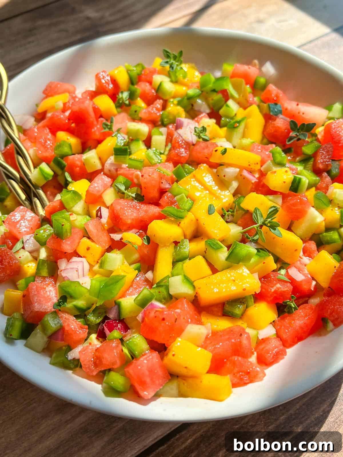 A bowl of colorful salsa made with diced watermelon, mango, green bell pepper, red onion, and fresh herbs, sitting on a wooden table in natural sunlight.