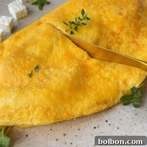 A golden-yellow Feta and Potato Omelet garnished with fresh herbs is served on a speckled plate, with cubes of white cheese and green sprigs on the side. A gold fork rests on the omelet.