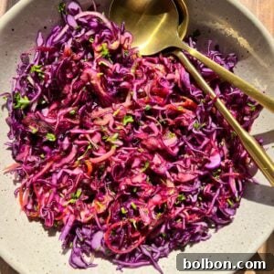 A bowl of colorful red cabbage coleslaw garnished with parsley is served in a light-colored dish. Two gold-colored utensils are placed on the side. The dish is set on a wooden surface, illuminated by natural light.