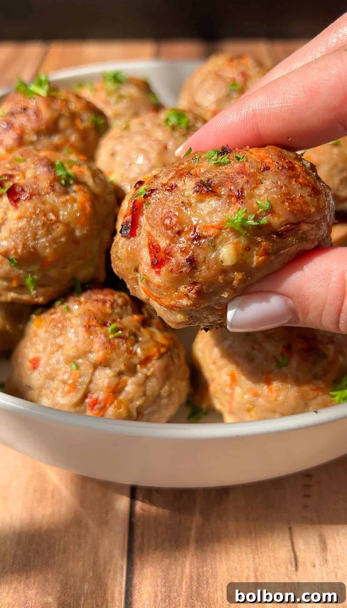 Close-up of a hand holding a meatball above a bowl filled with more meatballs. The meatballs are golden brown with small red and green flecks, garnished with chopped herbs. They are set on a wooden surface.