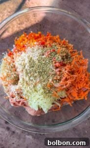 A bowl filled with minced meat, grated carrots, finely chopped red and orange bell peppers, and grated onions. The mixture is topped with various spices and seasonings, likely including herbs and ground pepper.