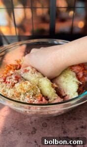 A hand mixes ingredients in a clear glass bowl. The mixture includes ground meat, chopped vegetables, and breadcrumbs, suggesting preparation for a dish like meatloaf or meatballs. Subtle light and shadow play across the scene.