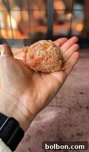 A person holds a raw meatball mixture with bits of vegetables like corn and carrots in their palm. They are wearing a smartwatch on their wrist. The background is softly blurred with warm lighting.