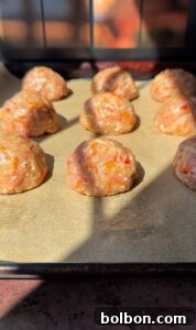 Raw meatballs with visible vegetable pieces are placed on parchment paper on a baking tray, illuminated by sunlight.