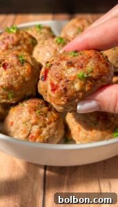 A hand holding a cooked meatball above a bowl filled with several meatballs, garnished with chopped parsley. The meatballs have a golden-brown exterior with visible herbs and spices. They are placed on a wooden surface.