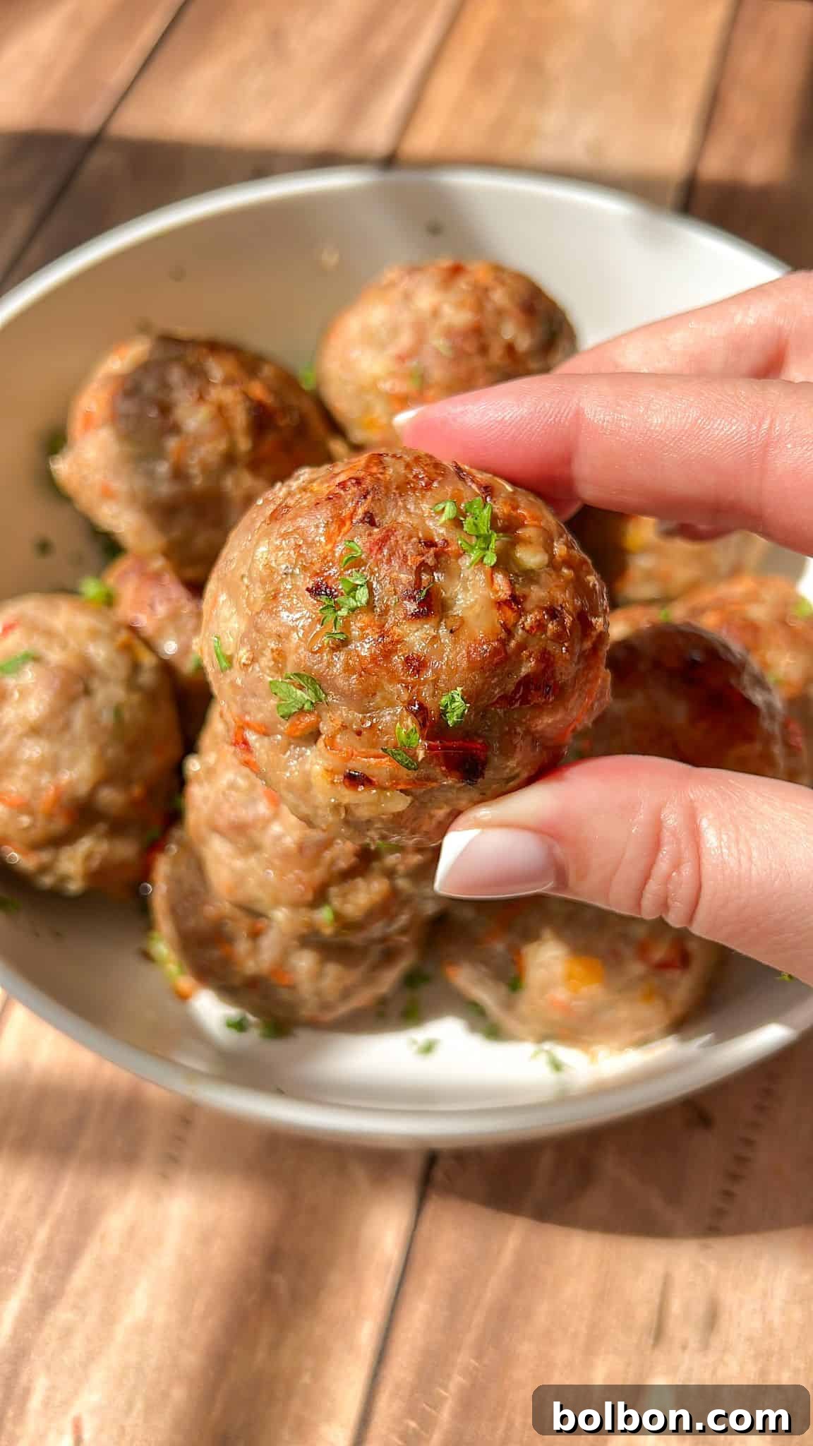 Close-up of a hand holding a cooked meatball garnished with herbs above a bowl filled with more meatballs. The meatballs are on a wooden table, basking in warm natural light.
