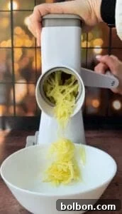 A person uses a white hand-crank grater to shred potatoes into a white bowl. The background is out of focus with warm, festive lights.