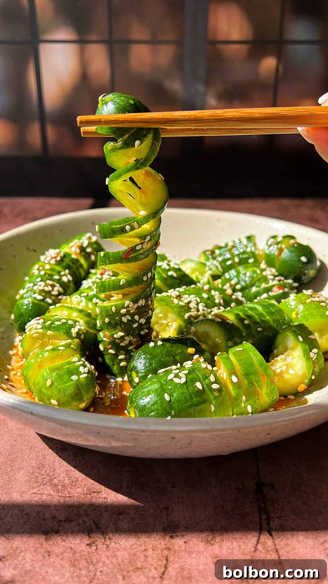 Refreshing Asian Cucumber Crunch 2 A bowl of spiral-cut cucumbers with a glossy, seasoned dressing, sprinkled with sesame seeds. A hand uses chopsticks to lift a spiral cucumber slice. Background features a softly lit window with grid patterns.