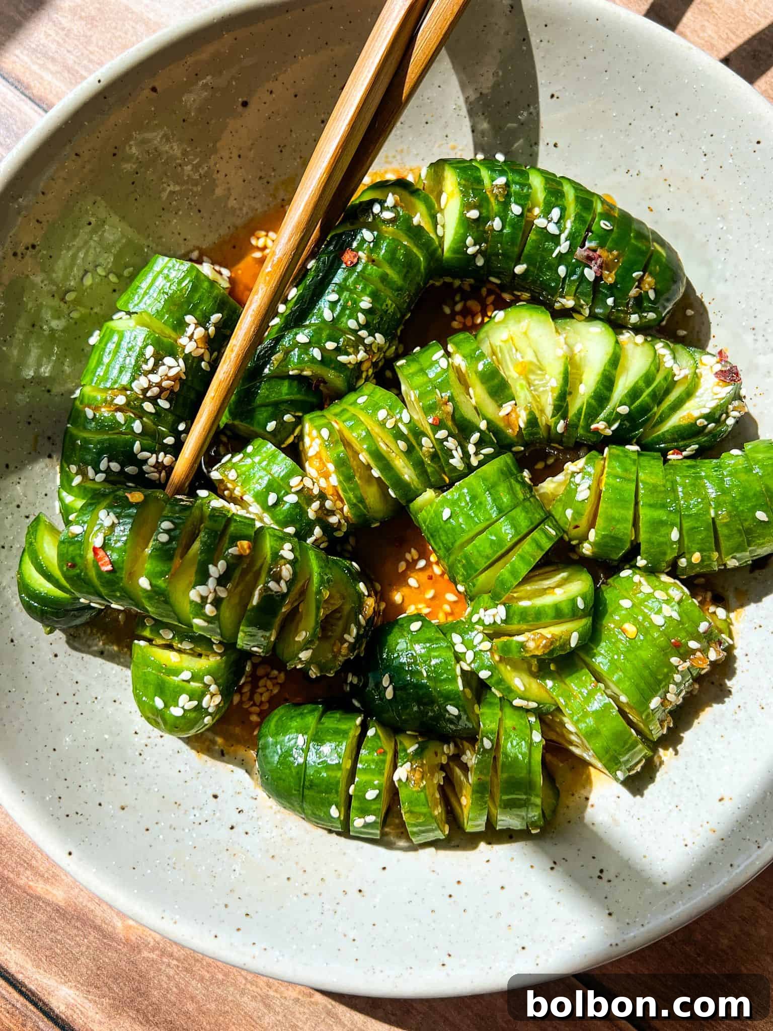 Refreshing Asian Cucumber Crunch 3 A plate of sliced cucumbers arranged in an S-shape, seasoned with sesame seeds, chili flakes, and a savory sauce. Chopsticks rest on the plate. The dish is bathed in sunlight on a wooden table.