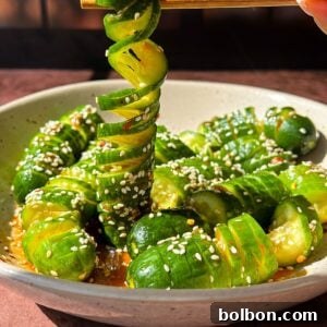A bowl of spiral-cut cucumbers with a glossy, seasoned dressing, sprinkled with sesame seeds. A hand uses chopsticks to lift a spiral cucumber slice. Background features a softly lit window with grid patterns.