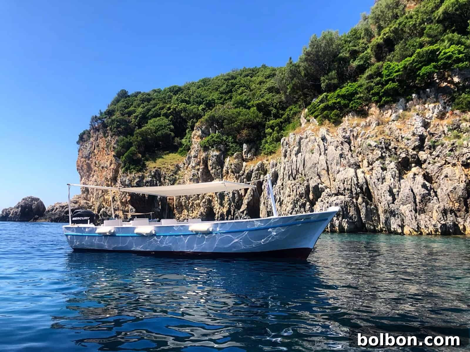 Effortless Greek Delights 3 A serene coastal scene features a boat with a beige canopy gently floating on crystal-clear blue water. Rocky cliffs, adorned with lush green vegetation, frame the tranquil inlet under a clear, azure sky. This picturesque setting in Greece is where I discovered the joy of Easy Greek Recipes.