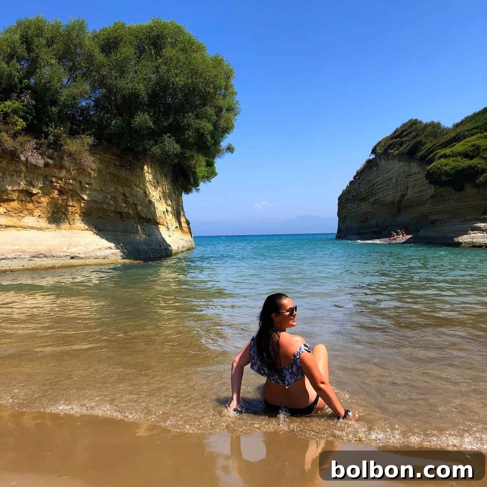 Effortless Greek Delights 4 A woman in a bikini relaxes on a sandy beach, gazing out at the calm blue sea within a rocky cove. Cliffs with lush green foliage frame the narrow inlet. The bright, sunny day and clear skies create a perfect, tranquil coastal scene, embodying the spirit of Greek relaxation and healthy living.