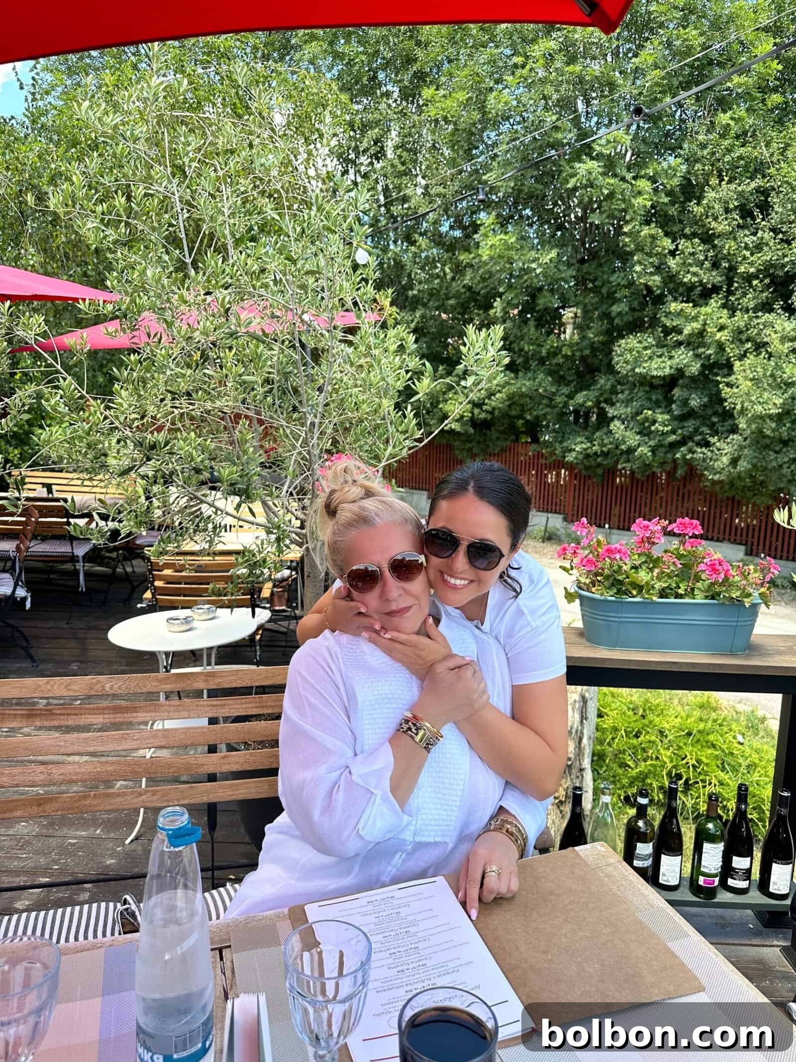 Two women wearing sunglasses sit together at an outdoor restaurant table before their weight loss journey. One woman hugs the other from behind. The table has menus, glasses, a water bottle, and a wine bottle nearby. The background features green trees and pink flowers.