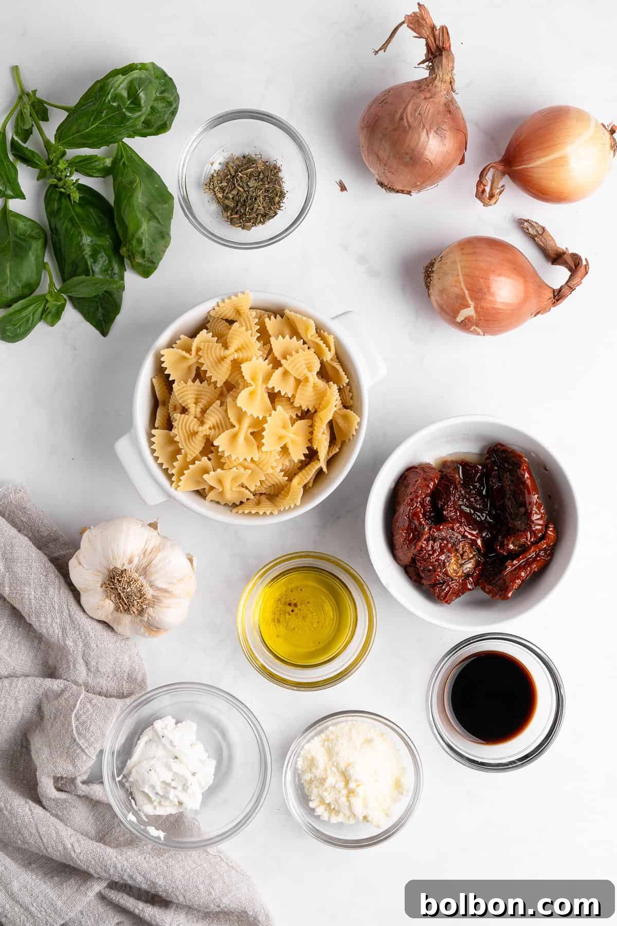 A marble countertop displays ingredients for easy Italian recipes, including uncooked bowtie pasta, fresh basil, dried herbs, whole onions, sun-dried tomatoes, a garlic bulb, olive oil, balsamic vinegar, ricotta cheese, and grated Parmesan cheese.