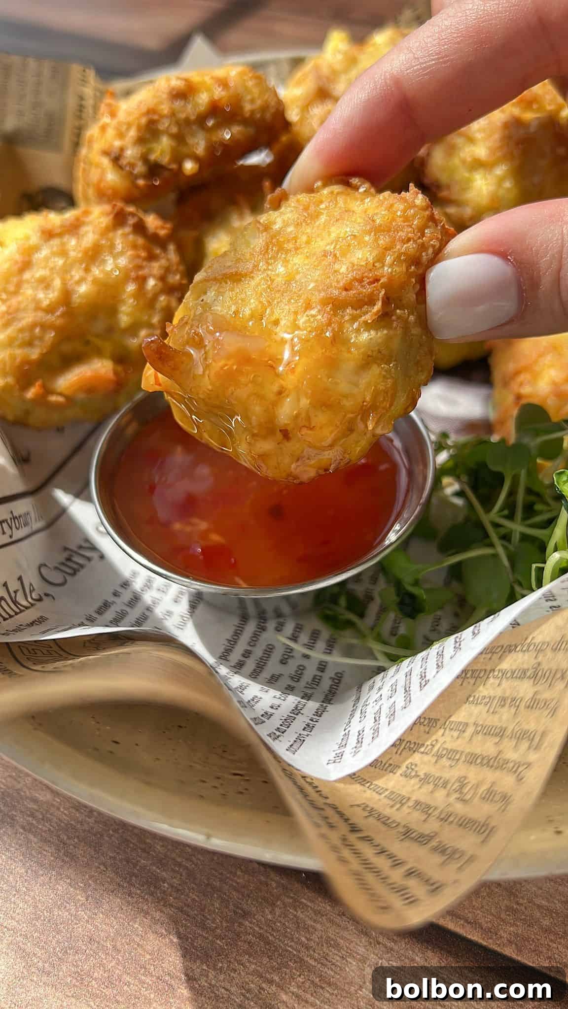 A hand holding a golden, crispy nugget dipped in orange sweet chili sauce, with more nuggets and a small dish of sauce on a newspaper-lined plate in the background.