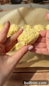 A person holds a rectangular, uncooked snack that appears to be made from instant ramen noodles; more similar snacks rest on a parchment-lined baking sheet in the background.
