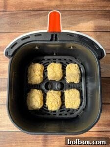 Six breaded nuggets are arranged in the basket of a black air fryer with a red handle, set on a wooden surface.