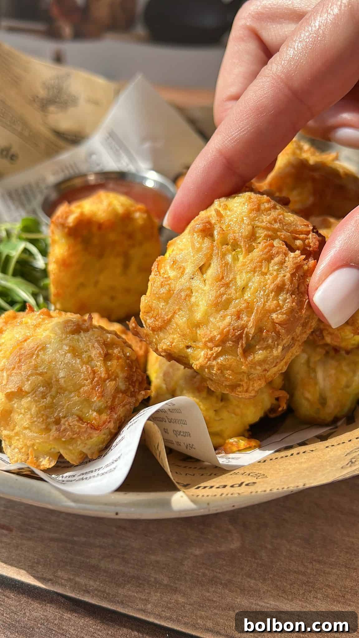 A hand with white nail polish holds a golden, crispy air fryer chicken nuggets above a bowl filled with more tater tots, a side of greens, and a cup of dipping sauce, all served on newspaper-like paper.