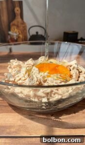 A glass bowl filled with shredded chicken and a raw egg yolk being poured over it, set on a wooden surface with kitchen items blurred in the background.