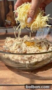 A hand sprinkles shredded cheese into a glass bowl containing shredded chicken and cracked eggs, all set on a wooden surface in bright natural light.