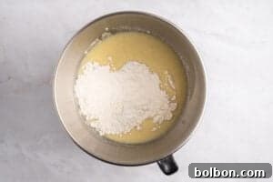 A metal mixing bowl containing flour and a yellowish liquid mixture, sitting on a light-colored marble surface.