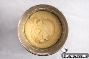 A metal mixing bowl filled with smooth, pale yellow cake batter sits on a white marble surface.