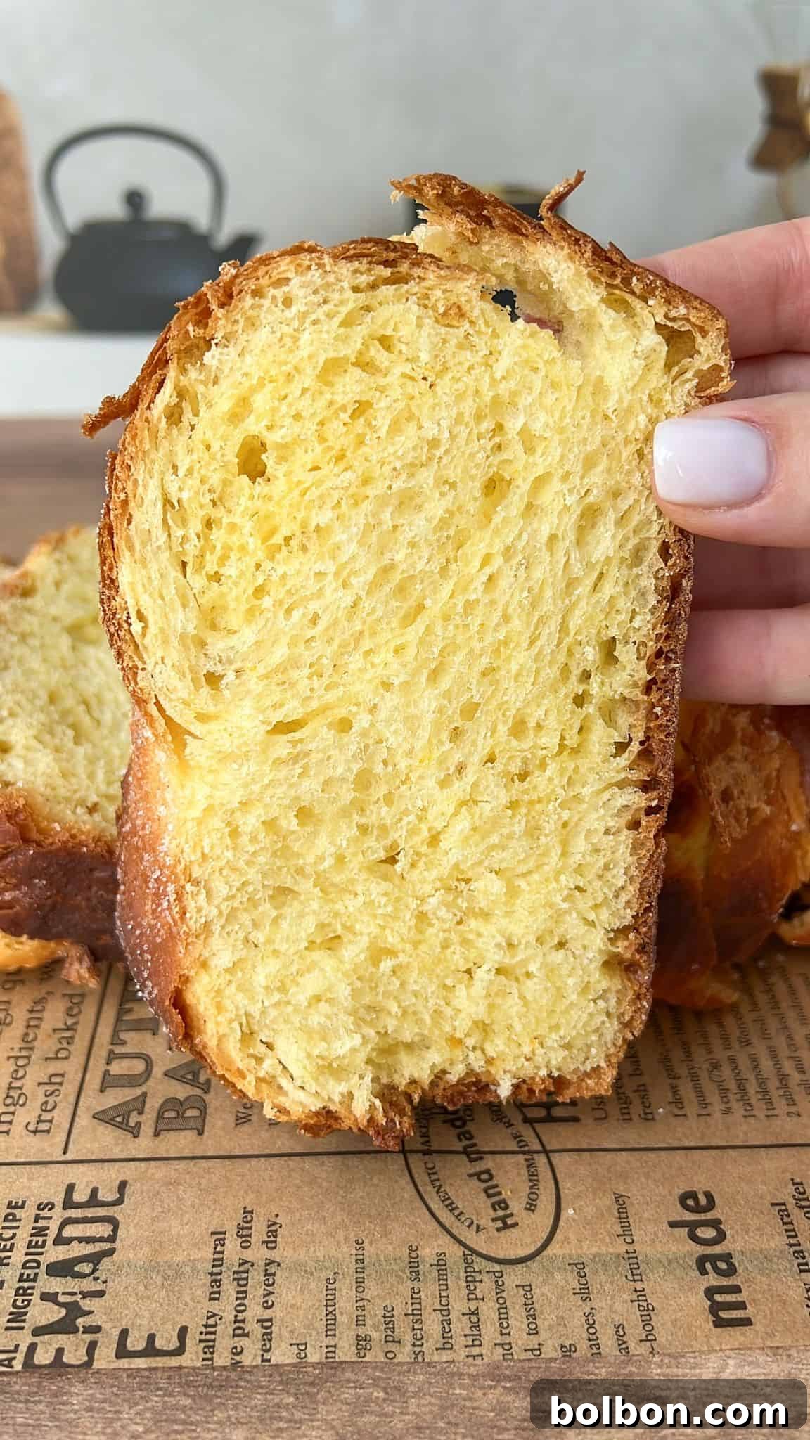 A hand holding a thick slice of golden, fluffy brioche bread with a soft, airy texture. A loaf and more slices are visible in the background on a rustic paper-lined wooden surface.