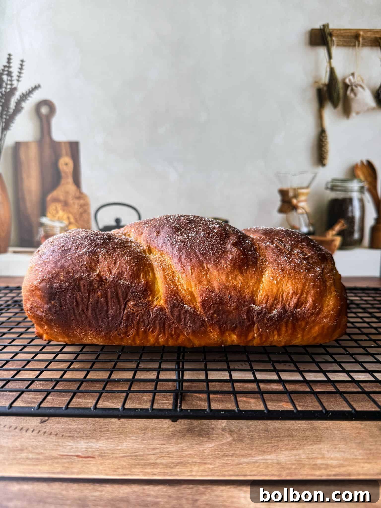 A golden-brown loaf of bread with a sugar-dusted top cools on a black wire rack in a cozy kitchen setting with wooden utensils and jars in the background.