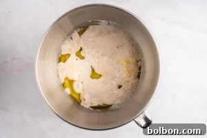 A metal mixing bowl containing flour, yeast, oil, and water, with ingredients partially mixed, placed on a white marble countertop.