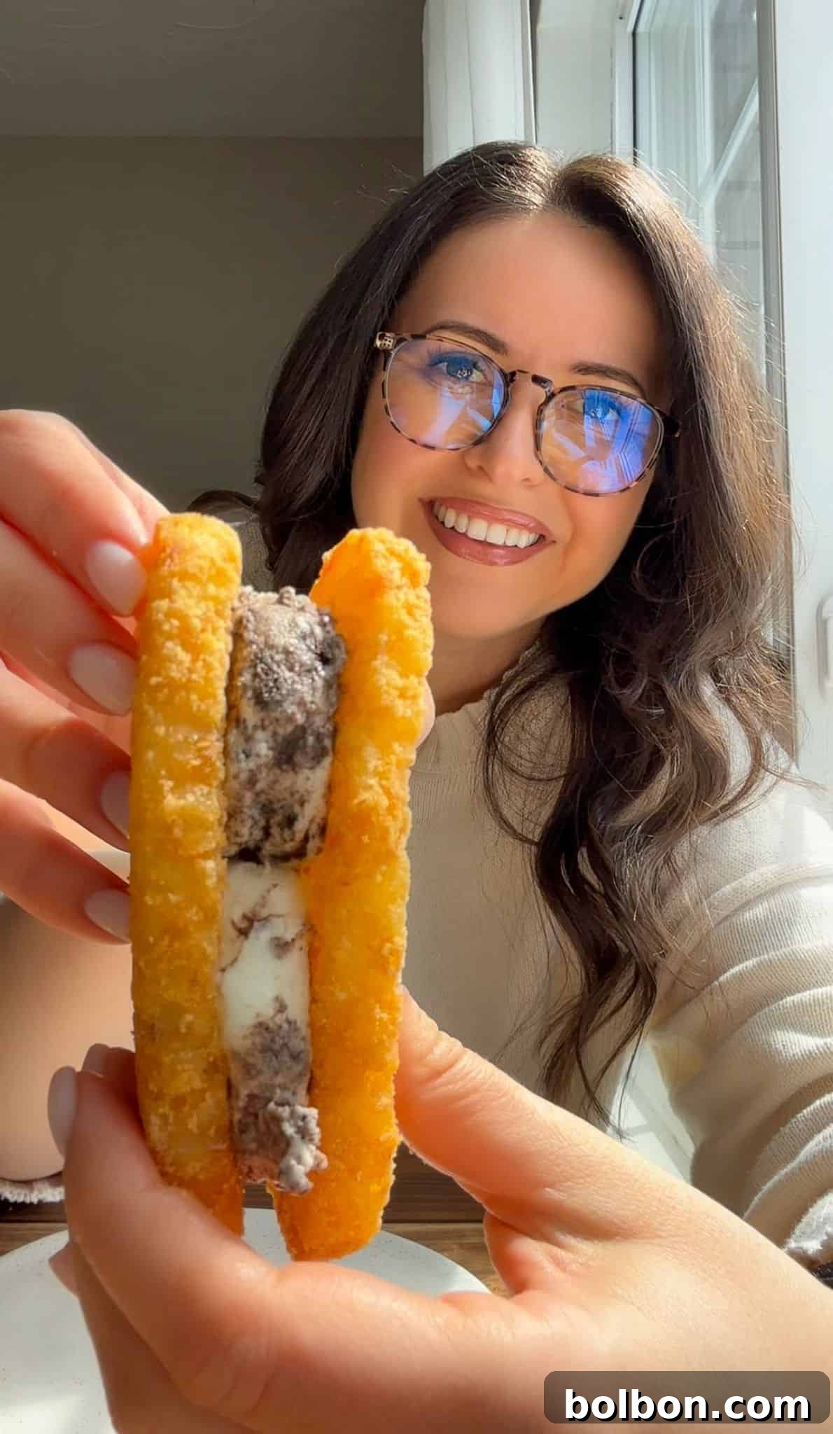 A smiling woman with long dark hair and glasses holds up a dessert made of two hash browns with ice cream sandwiched between them. Sunlight shines through a window behind her.
