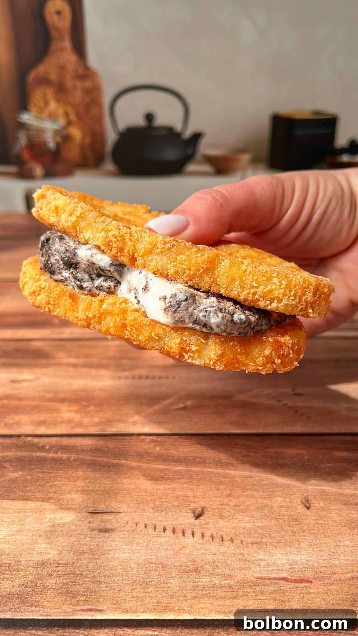 A hand holds a sandwich made with two crispy, breaded hash browns as buns, filled with cookies and cream ice cream, against a wooden table background with kitchen items blurred in the distance.