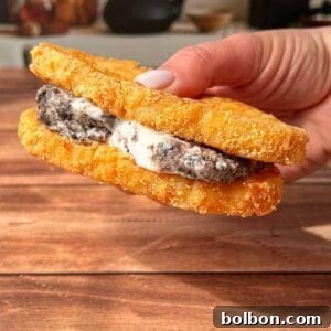 A hand holds a sandwich made with two crispy, breaded hash browns as buns, filled with cookies and cream ice cream, against a wooden table background with kitchen items blurred in the distance.