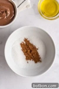 A white bowl filled with granulated sugar and cocoa powder sits on a marble surface, ready for coating delicious French Toast Rollups, alongside a bowl of chocolate spread and a small bowl of yellow oil.