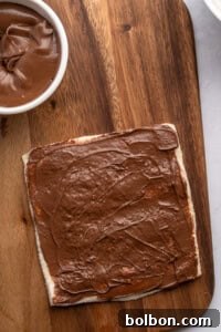 A slice of bread spread with chocolate hazelnut spread sits on a wooden cutting board, next to a small white bowl of more spread—perfect for making delicious French Toast Rollups.