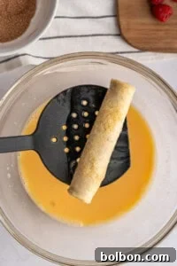 A rolled tortilla for French Toast Rollups is held over a bowl of beaten eggs with a black slotted spatula, ready to be coated. In the background, cinnamon sugar awaits alongside a wooden board topped with strawberries.
