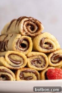A stack of French Toast Rollups filled with chocolate and drizzled with more chocolate sits on a white plate next to a fresh raspberry.