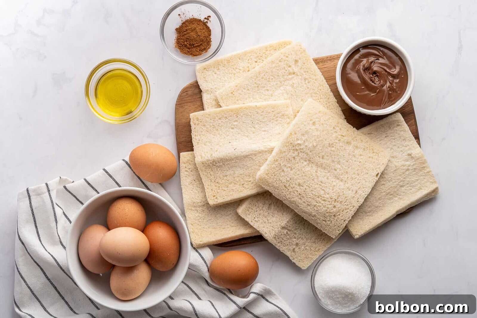 Several slices of white bread on a cutting board, surrounded by a bowl of eggs, a small bowl of chocolate spread, a bowl of oil, a dish with cinnamon, and a bowl of sugar on a white surface with a striped cloth.