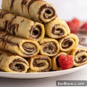 A stack of rolled crepes, reminiscent of French Toast Rollups, filled with chocolate hazelnut spread and drizzled with more chocolate, is arranged on a white plate with fresh raspberries scattered around.