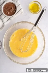 A glass bowl with whisked eggs and a metal whisk, surrounded by bowls containing olive oil, chocolate spread, and sugar—perfect ingredients for making delicious French Toast Rollups on a white countertop.